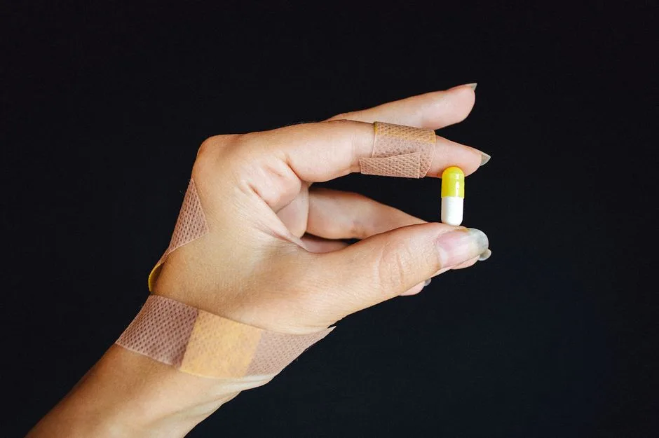 A hand with bandages holding a yellow and white capsule against a black background.