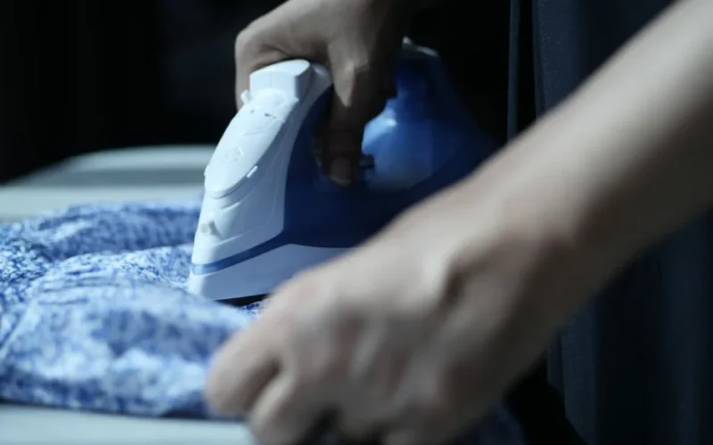 Detailed view of hands using an electric iron on blue fabric indoors.