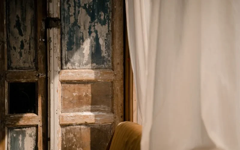 Sunlit rustic wooden door with aged texture partially covered by flowing curtains.