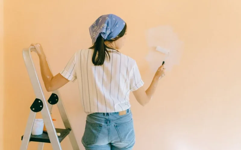 A woman with a bandana using a paint roller to paint a peach-colored wall indoors.