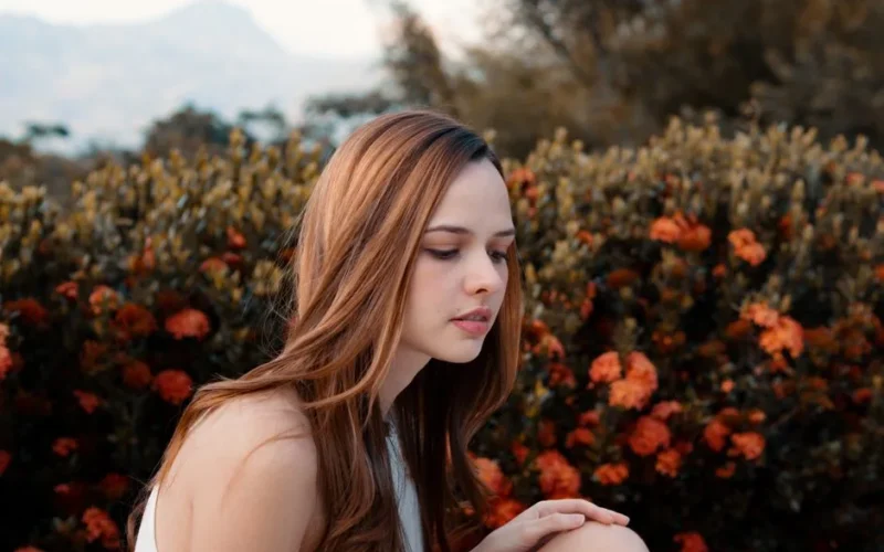 A young woman with long brown hair poses gracefully in front of vibrant orange flowers outdoors.