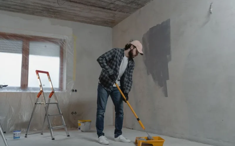 A man painting a room with a roller, using a stepladder, wearing casual attire. Renovation scene.