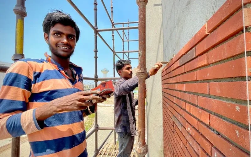 Two construction workers laying red bricks on scaffolding outside a building.