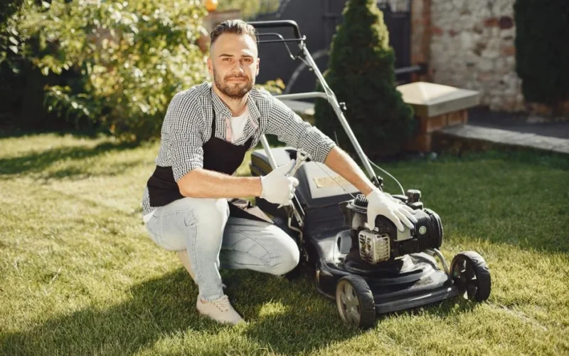 Adult man wearing apron and gloves maintains a lawn mower on a sunny day.