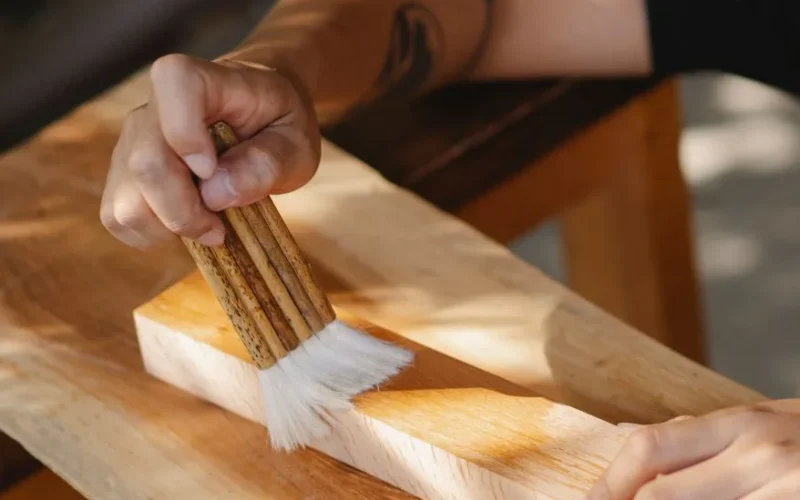 High angle of unrecognizable artisan using brush for varnishing wooden beam while working in professional workshop
