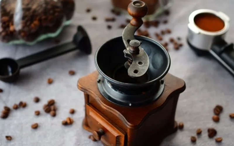 Vintage manual coffee grinder surrounded by scattered coffee beans and tools on a rustic table.