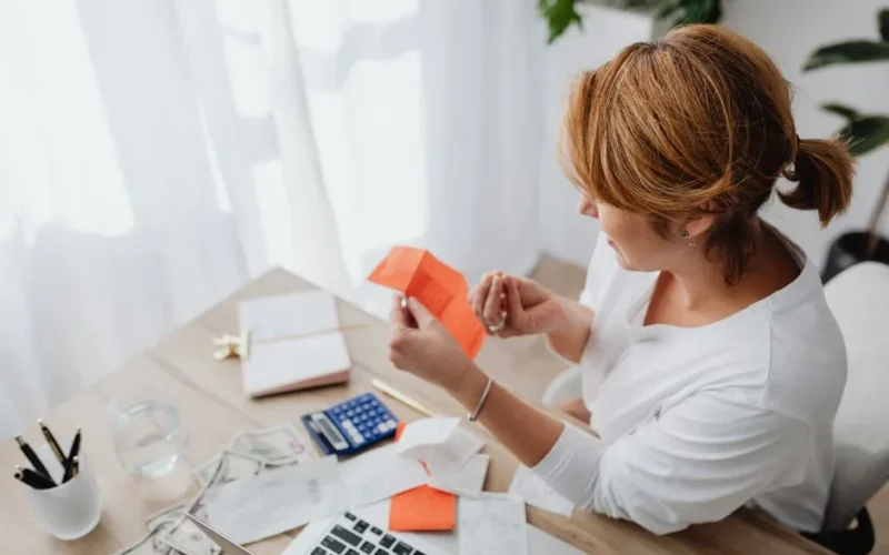A woman organizing her finances with bills and receipts at a home office desk using a calculator and laptop.