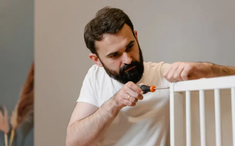 A man focusing closely on assembling a baby crib with a screwdriver in a well-lit room.