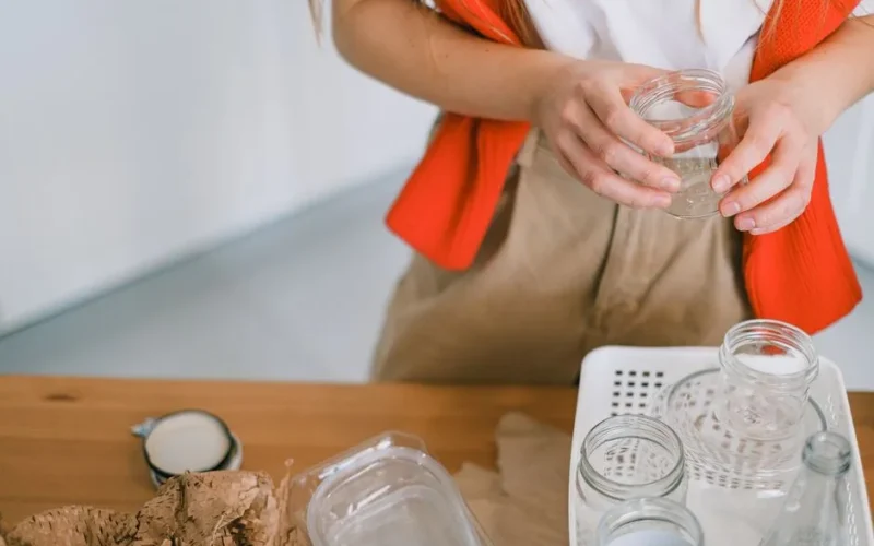 Crop anonymous female in casual clothes standing with glass jar near table while sorting out paper plastic and glass rubbish for recycling