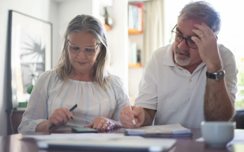 Senior couple calculating expenses at home office desk with documents and notes.