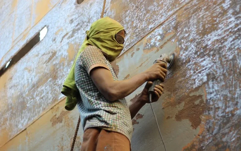 A worker using a grinder to polish rusted metal, wearing protective clothing outdoors.