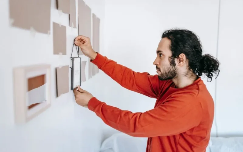 A man in a red sweater hangs a picture frame on a white wall in a modern room.