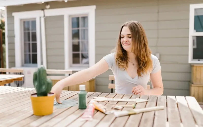 Young woman crafting outdoors with paints and brushes on a wooden table.