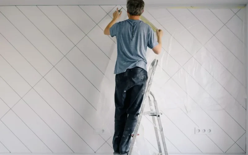 Full body back view of faceless male in shirt and dirty pants repainting wall while standing on ladder in bright empty room in daytime
