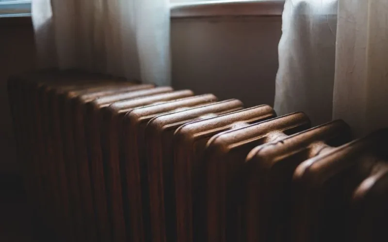 Aged heating battery made of cast iron near windowsill with curtains in house