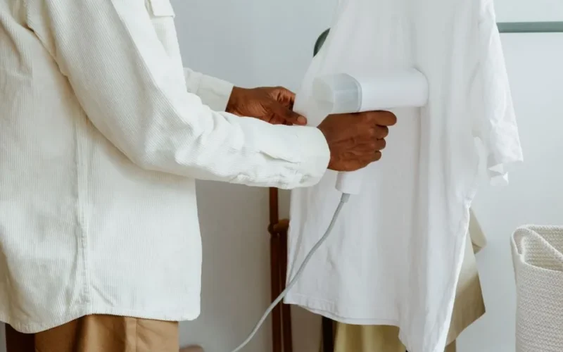 A man uses a steam iron to remove wrinkles from a white shirt hanging on a rack indoors.