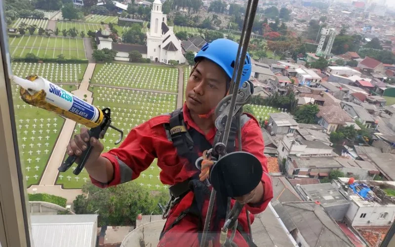 A professional window cleaner in Jakarta using safety gear and sealant with a cityscape vista backdrop.