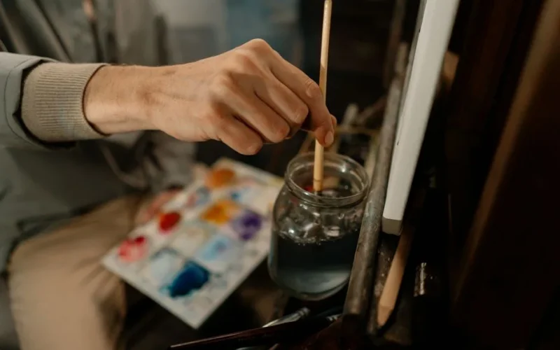 Close-up of an artist's hand dipping a paintbrush in a water jar with paint palette nearby.