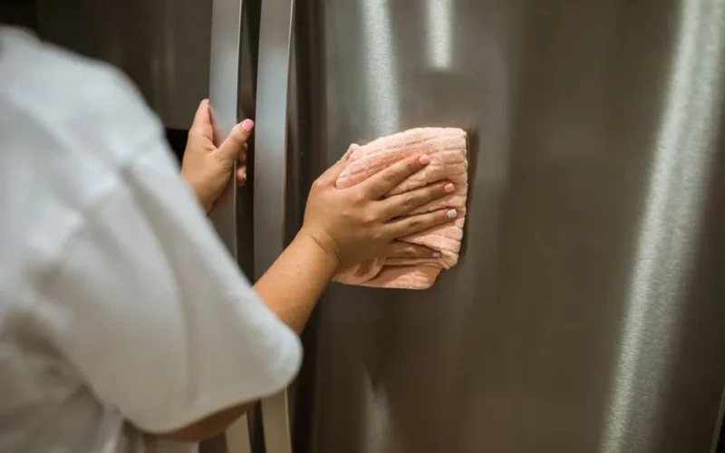 Hands using cloth to wipe stainless steel refrigerator. Indoor cleaning action.