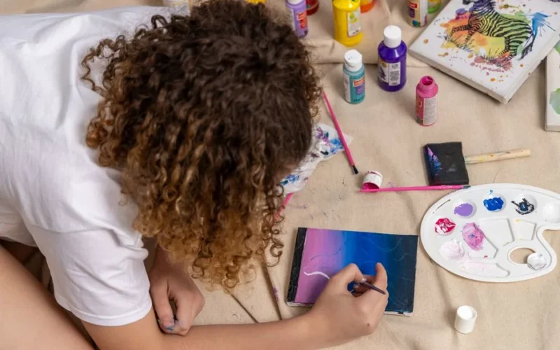 A young artist paints with acrylics on a canvas surrounded by supplies, showcasing creativity.