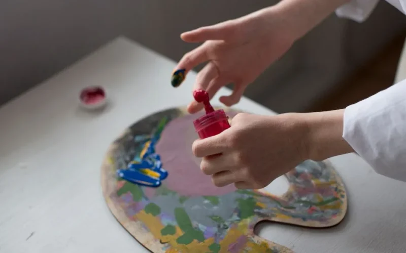 Close-up of an artist's hands mixing colorful paint on a palette indoors.