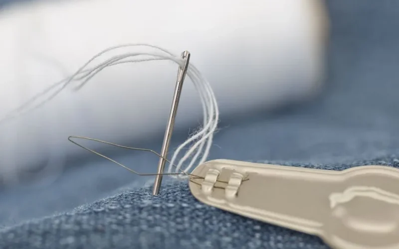 Detailed view of a sewing needle with white thread on blue denim fabric with needle threader.
