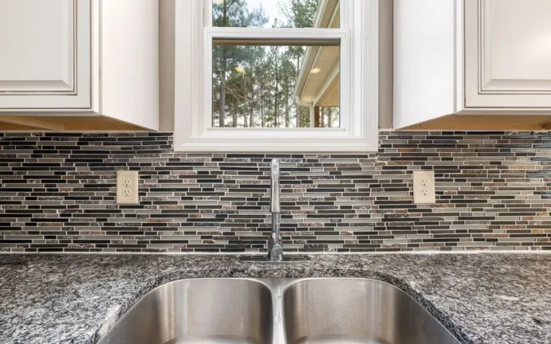 Close-up of a stainless steel sink on a granite countertop with a tiled backsplash and a window view.