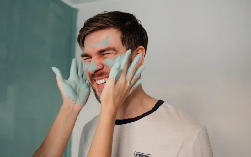 Fun indoor scene of a man laughing while getting his face painted with blue paint.
