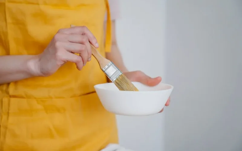 Close-up of a woman in a yellow apron mixing paint in a white bowl with a brush.