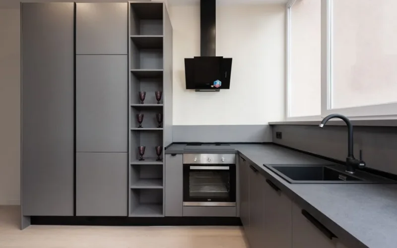 Interior of light gray kitchen corner furnished with cupboards and stone counter with built in oven and sink placed with big windows in apartment