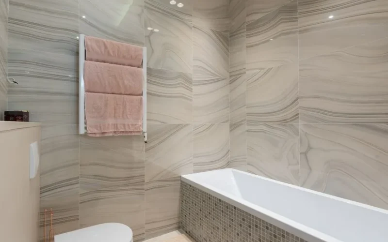 Contemporary bathroom interior with toilet bowl against bathtub and tiled walls with towels on rack in light house