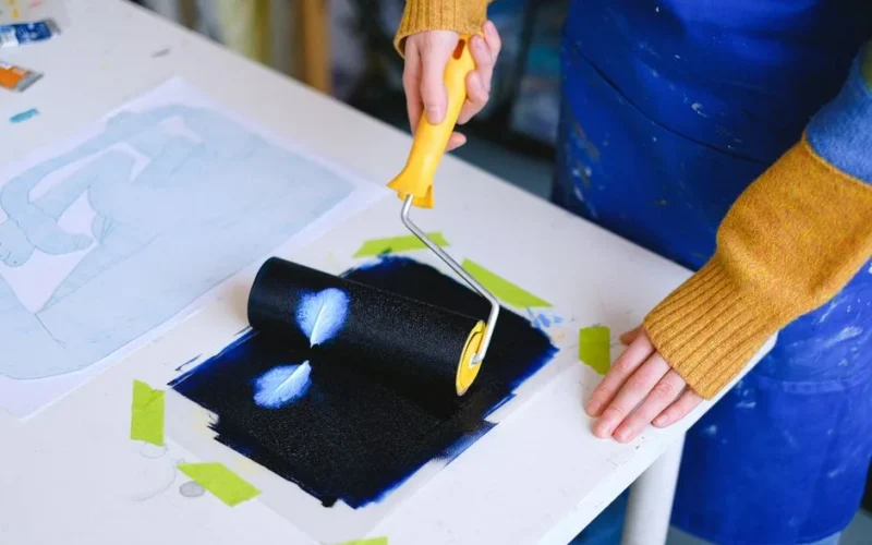 High angle of crop anonymous female artist in casual outfit and dirty apron standing near table in light studio while rolling on paper with roller covered in paint