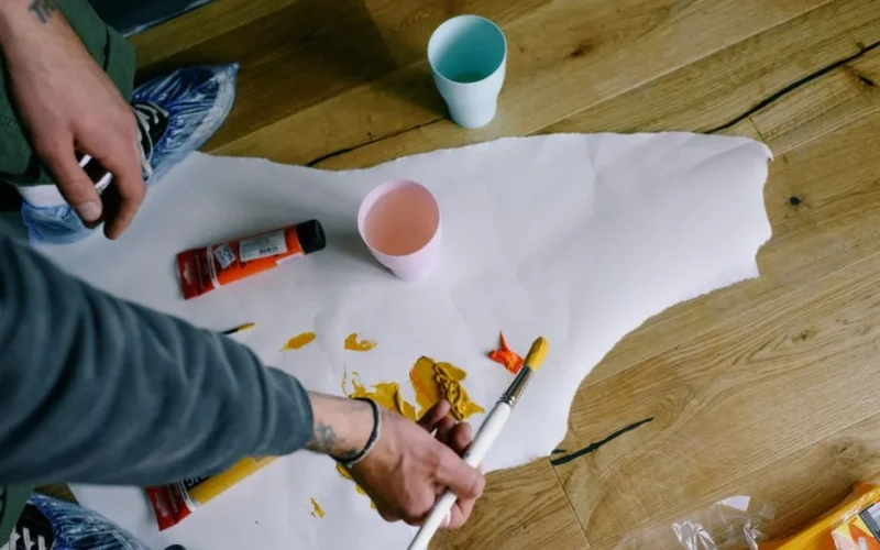 From above crop anonymous male artist in casual clothes and overshoes sopping paintbrush in yellow paint on paper snatch on floor