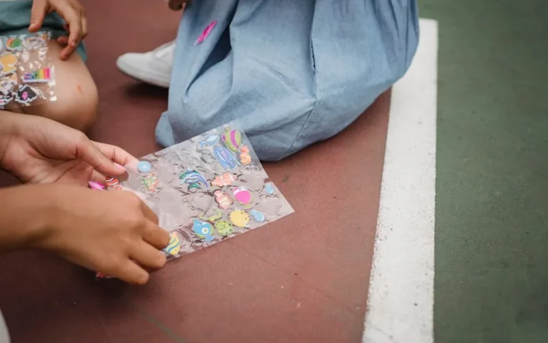 Close-up of children exchanging colorful stickers while sitting outdoors on a court.