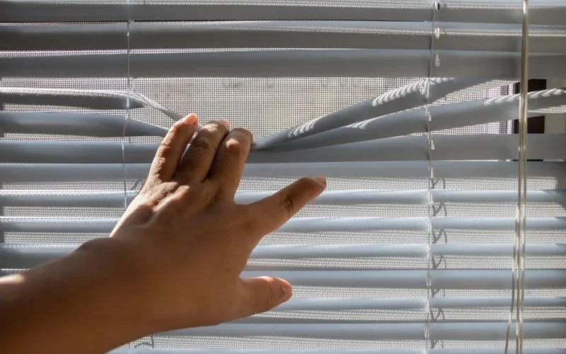 A hand gently opens window blinds, letting sunlight stream indoors.