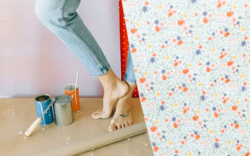 A woman decorates a room with floral wallpaper, surrounded by paint cans and a brush.