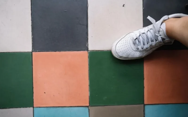 A white sneaker stepping on a colorful tiled floor featuring green, orange, and blue tiles.
