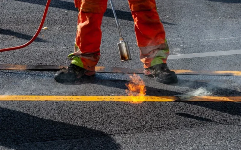 A construction worker using a torch to seal a road's asphalt surface with precision and care.
