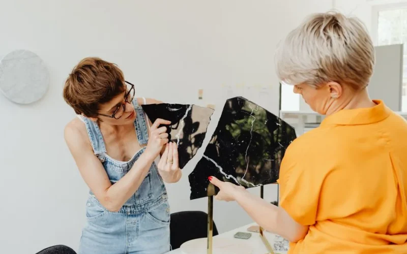 Two women examining broken marble tiles indoors, discussing repair strategies.