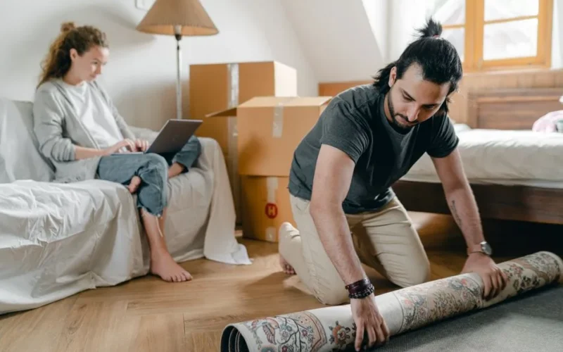 Barefoot girlfriend surfing internet on netbook while sitting on cozy sofa and ethnic man in smart watch laying rug on parquet after moving into new apartment