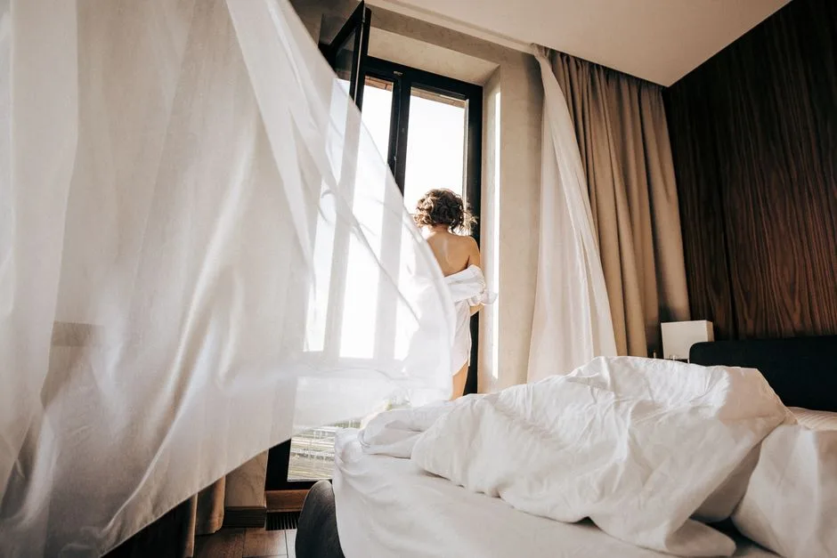 A person stands by an open window in a bedroom with flowing curtains and soft morning light.